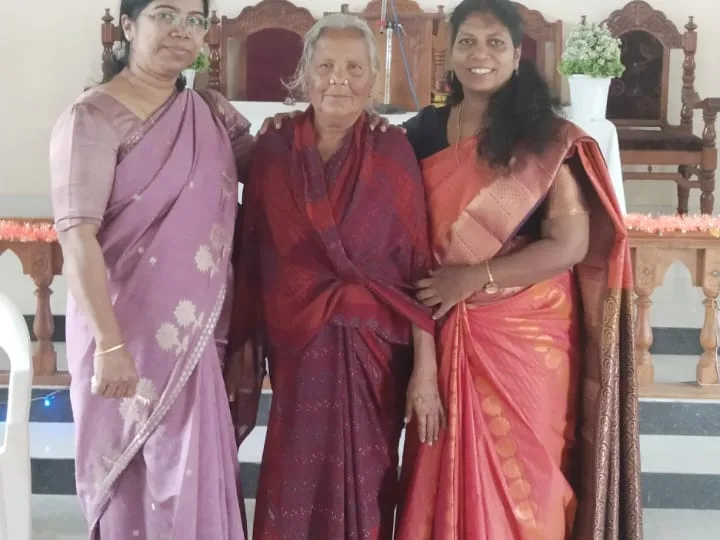 Three women standing together in front of a cross in a church, with an elderly woman in the middle