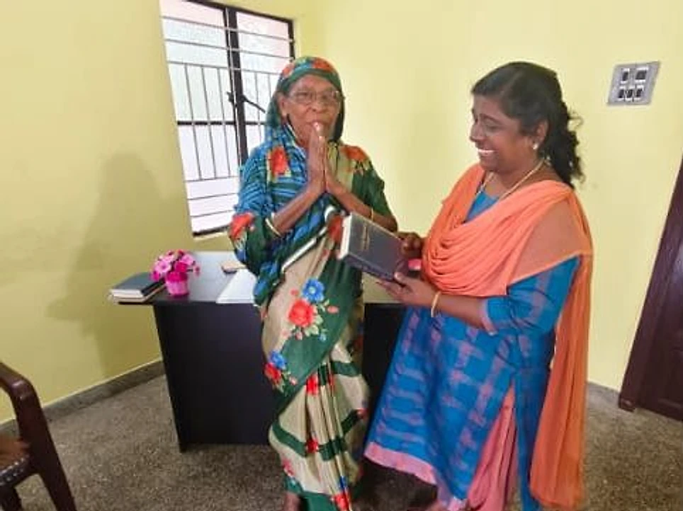 A woman in a blue outfit with an orange shawl holding a black book beside a smiling elderly woman with her hands pressed together