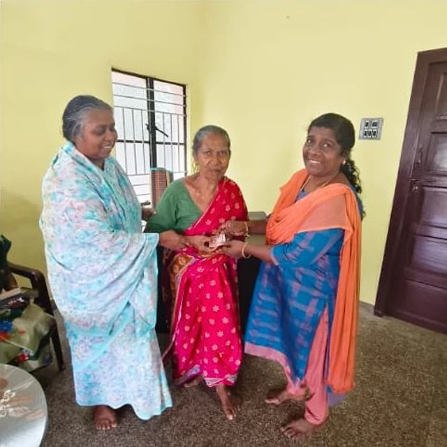 Three women standing in a room with yellow walls: a woman in a blue outfit with an orange shawl hands a small item to an elderly woman in a red saree, with another elderly woman in a light blue saree beside them