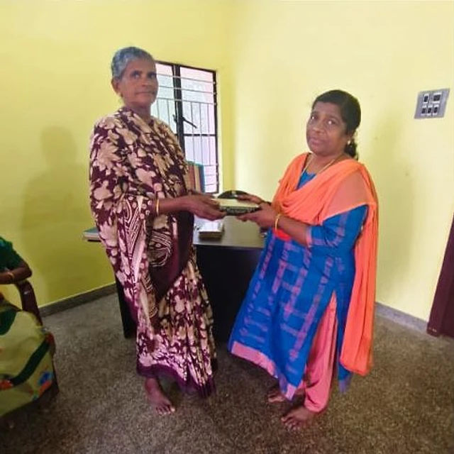 A woman in a blue outfit with an orange shawl handing a black book to an elderly woman in a patterned saree, in a room with yellow walls
