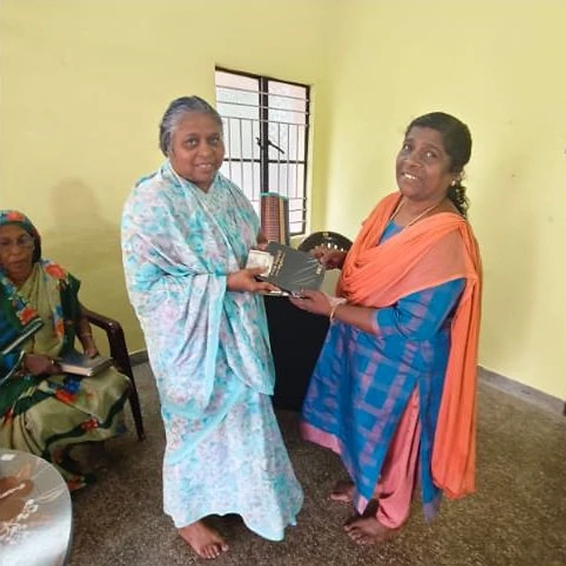 A woman in a blue outfit with an orange shawl and an elderly woman in a light blue saree smiling and holding a black book between them, with another elderly woman seated at the left