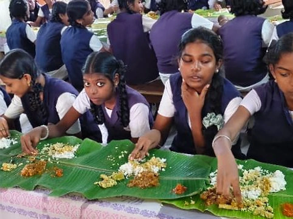 Schoolgirls in purple and white uniforms eating a meal from banana leaves at a classroom table