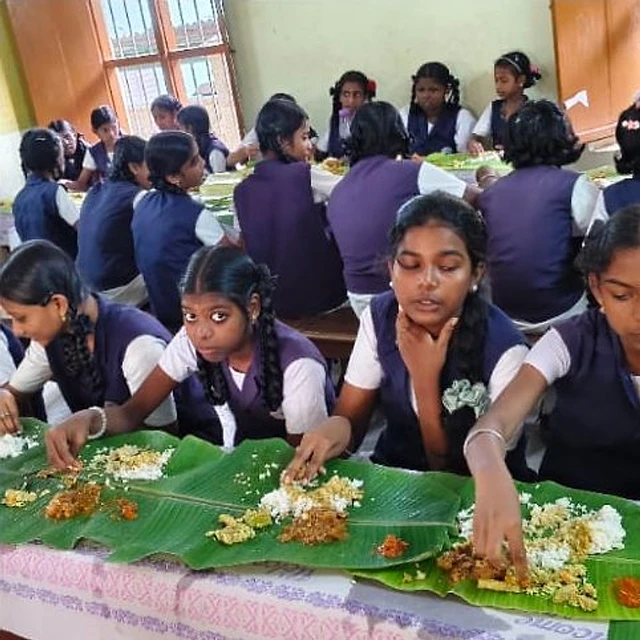 Schoolgirls in purple and white uniforms sitting at a classroom table and eating a meal from banana leaves