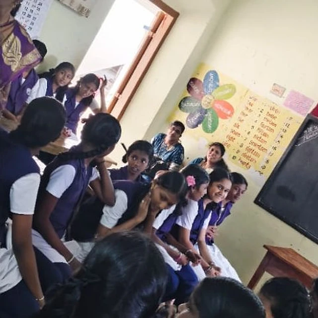 Schoolgirls in purple and white uniforms sitting close together in a classroom with a chart on the wall