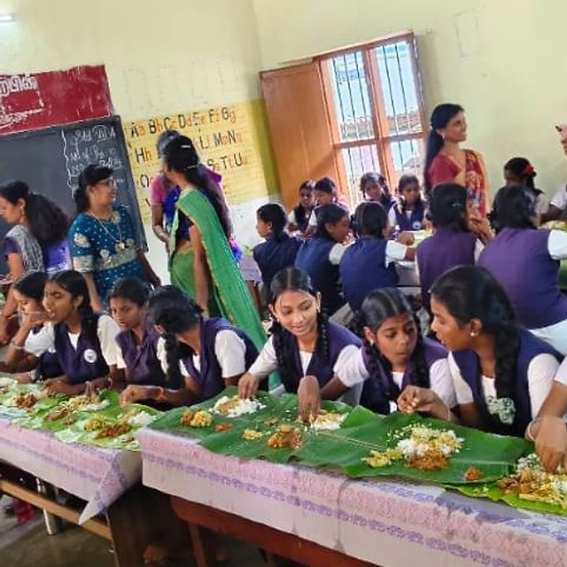 Rows of schoolgirls in purple and white uniforms eating a meal from banana leaves laid along long tables in a classroom, with women standing at the back