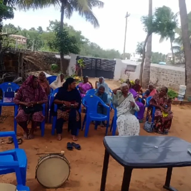 Women seated on blue plastic chairs in a yard under palm trees, with a drum on the ground beside a table