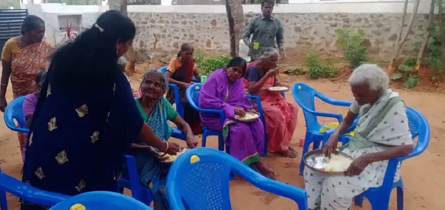 Women seated on blue plastic chairs in a yard being served food, with a woman in a dark blue saree bending to help