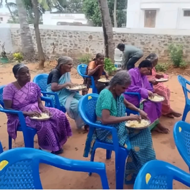 Women seated on blue plastic chairs eating rice from plates, in front of a stone wall