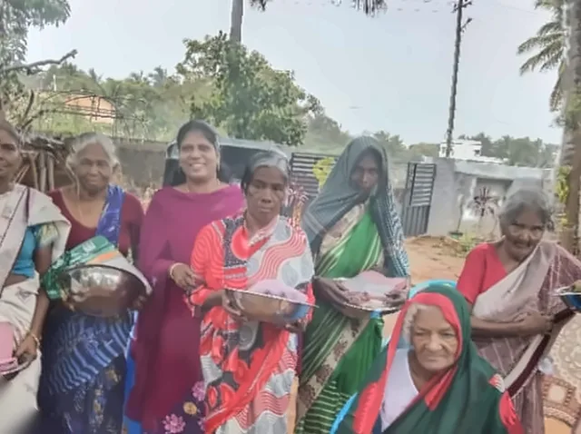 Women standing together holding steel bowls and bundles of cloth, with an elderly woman in a green and red headscarf seated in front