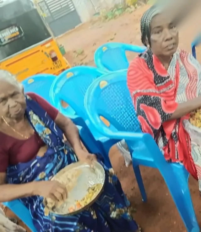 Two elderly women seated on blue plastic chairs eating food from plates, with a yellow auto-rickshaw behind them