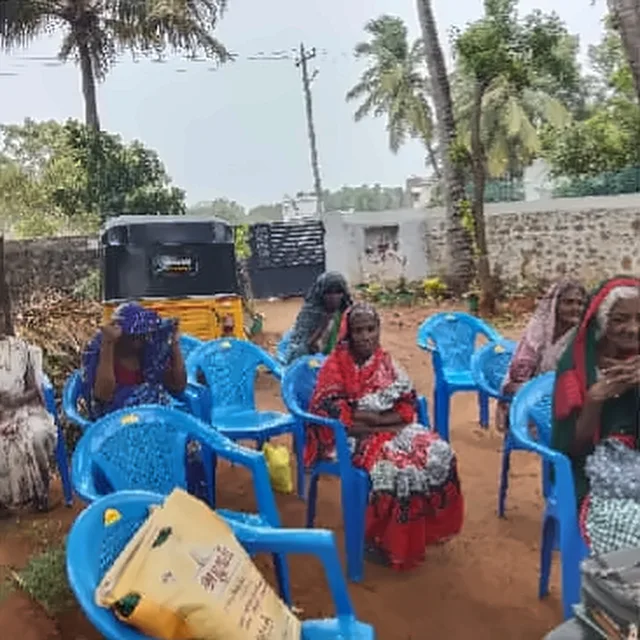 Elderly women seated on blue plastic chairs in a yard, with a yellow sack in front of them