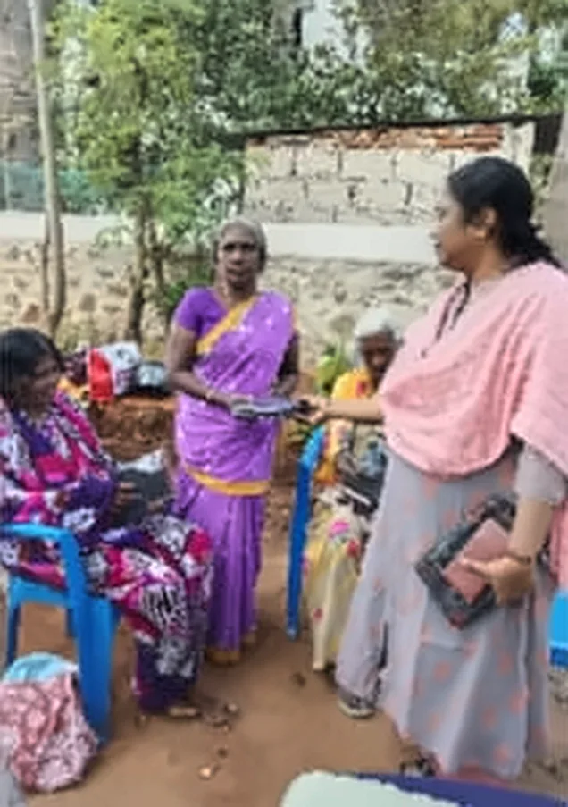 A woman in a pink shawl with a woman in a purple saree, with other women seated on chairs behind them