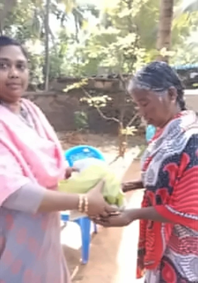 A woman in a pink shawl handing a yellow bag to an older woman in a red patterned saree