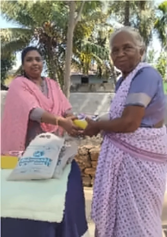 A woman in a pink shawl behind a table of bags, handing a gift to an older woman in a lilac saree