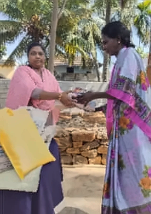 A woman in a pink shawl handing a gift to a woman in a blue and pink saree, with a yellow bag on the table