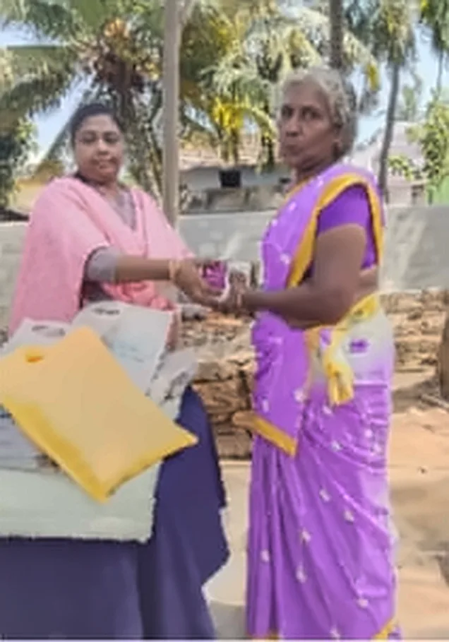 A woman in a pink shawl handing a gift to a woman in a purple saree with a yellow border