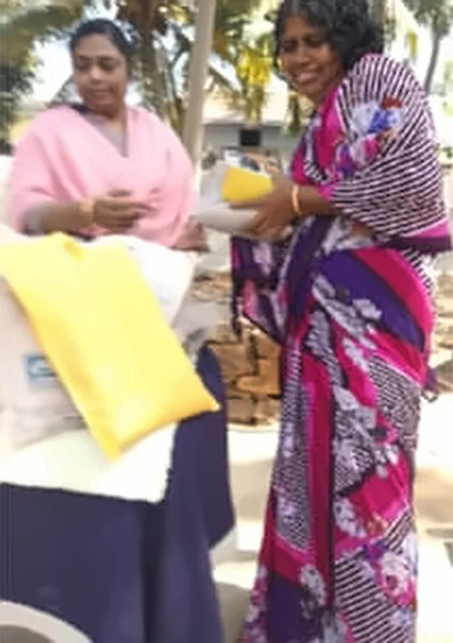 A woman in a pink shawl handing a gift to a woman in a purple and pink striped saree