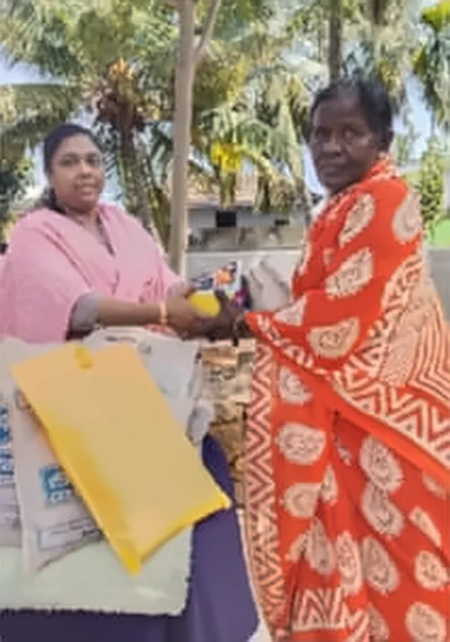 A woman in a pink shawl handing a gift to a woman in an orange patterned saree