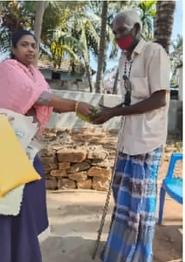 A woman in a pink shawl handing a gift to an older man in a face mask, who is leaning on a stick
