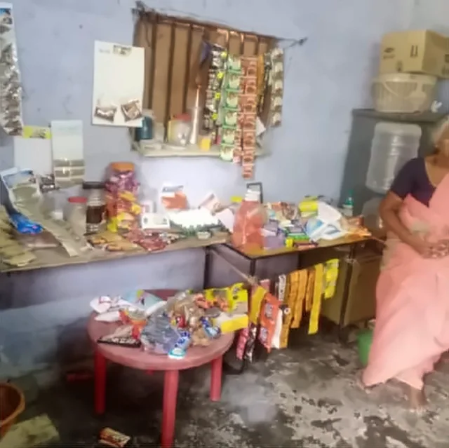 A small shop with goods on shelves, a table and a stool, and a woman in a pink saree seated at the side