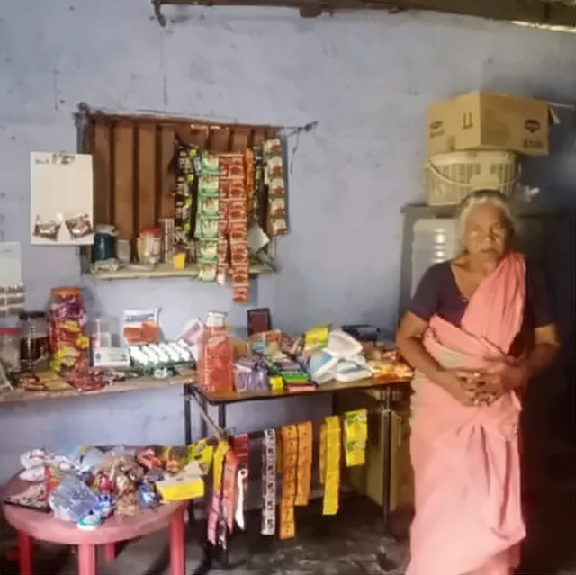 An elderly woman in a pink saree standing beside her small shop, with goods displayed on shelves and tables
