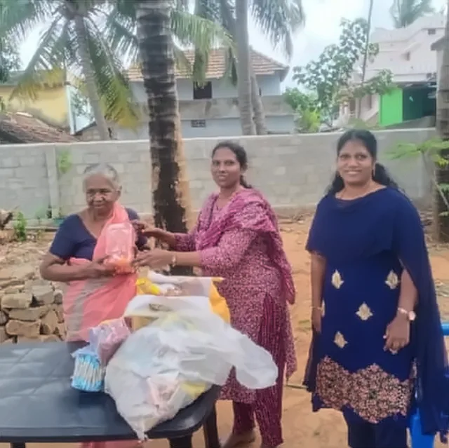 Two women giving bags of supplies to an elderly woman in a pink saree, outdoors in front of a stone wall