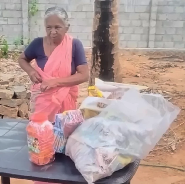 An elderly woman in a pink saree standing at a table with bags of supplies and a jar of sweets