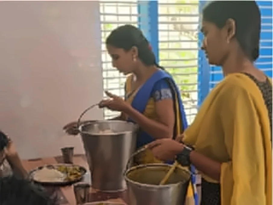 Two women serving food from steel vessels to a person seated at a table
