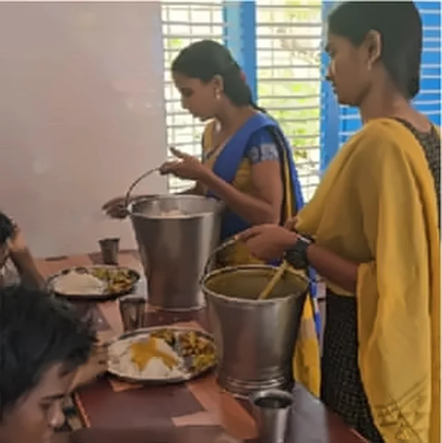 Two women serving food from steel vessels to a person sitting at a table