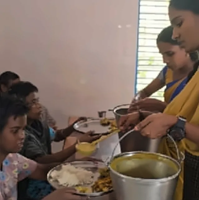 A woman serving rice and curry from a steel vessel to children seated at a table