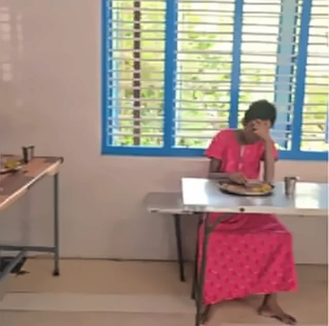 A woman in a pink dress sitting alone at a long table by a window with blue shutters