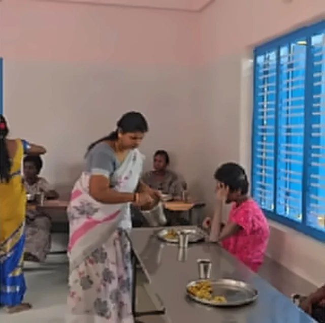 A woman in a white and pink saree serving food at a table, with a girl in a pink dress seated nearby