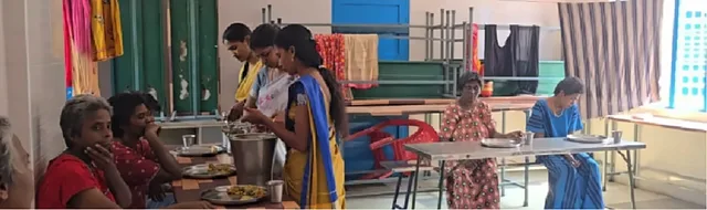 A wide view of a dining hall, with women serving food from a steel pot and others seated at tables with plates