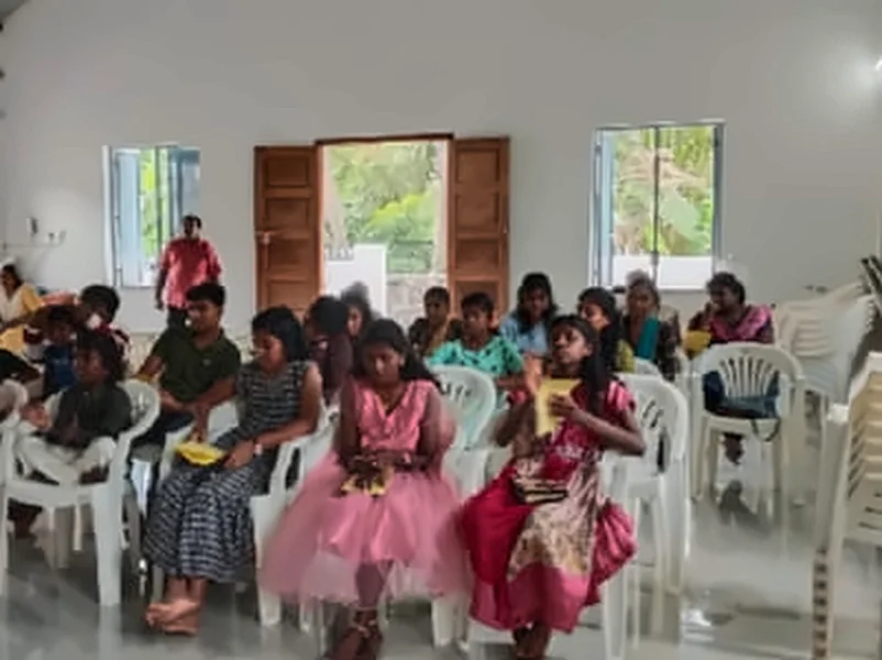 Children and teenagers seated on white plastic chairs in a hall, with two girls in pink dresses in the front row