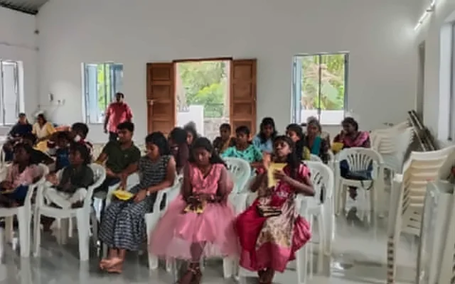 Children and teenagers seated in rows on white plastic chairs in a hall, with two girls in pink dresses in the front row
