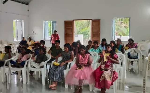 Children and teenagers seated in rows on white plastic chairs in a hall during the Bible school