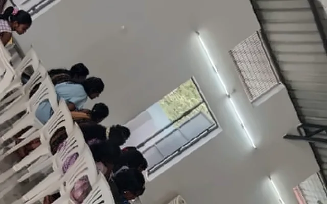 A tilted view of rows of children seated on white chairs in a hall
