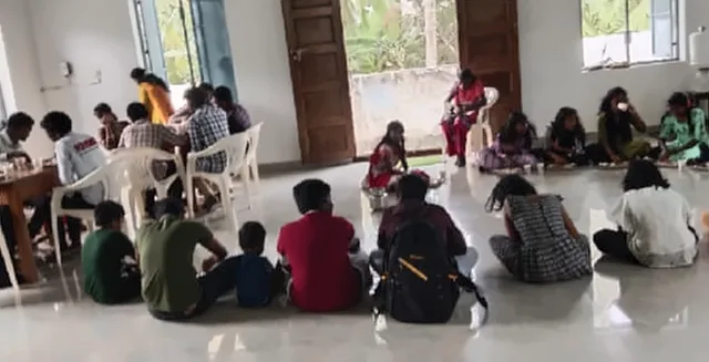 Children and helpers sitting on the floor of a hall, eating a meal
