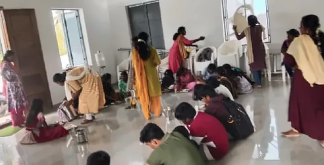 Children sitting on the floor of a hall eating, with women serving at the back