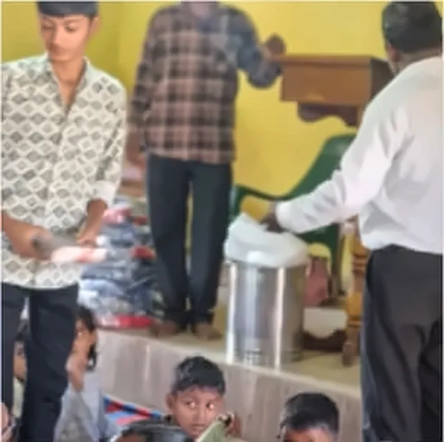 A boy in a patterned shirt standing near a man in a white shirt who is handing out books at a wooden stand, in a hall with yellow walls
