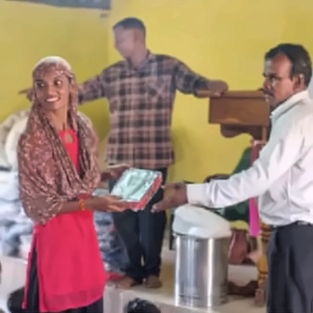 A smiling woman in a red top and a patterned headscarf receiving a Bible from a man in a white shirt