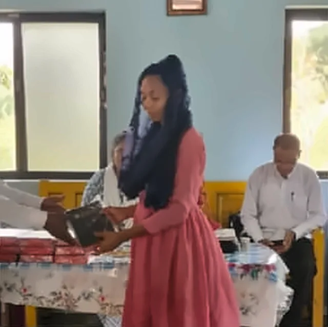 A girl in a pink dress receiving a Bible at a table with a flowered tablecloth, with an older man seated behind her