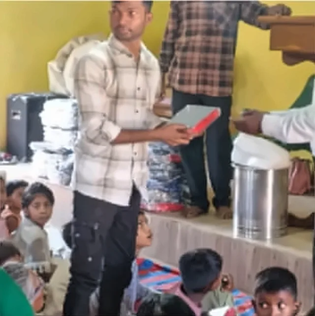 A man in a checked shirt giving out a Bible, with children seated on the floor in front of him