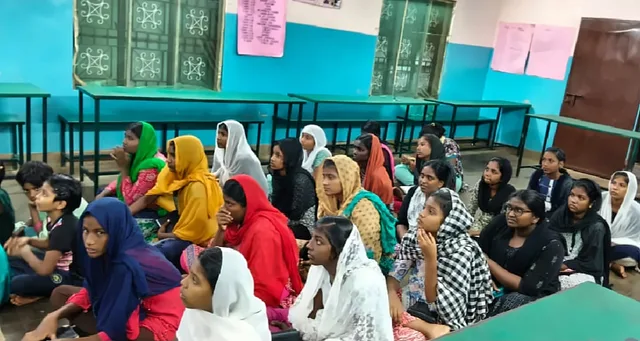 Girls in colourful headscarves sitting together on the floor of a hall with green benches, looking ahead