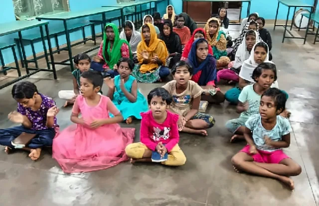 Girls and young children sitting together on the floor of a hall, clapping and singing