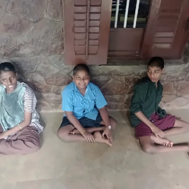 Three young people sitting on the floor in front of a stone wall, beneath a wooden window