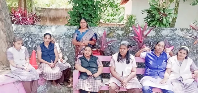 A group of girls in uniforms sitting on a pink bench in a garden, with a woman in a blue saree behind them
