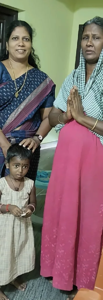 A woman in a blue saree standing with a woman in a pink dress, who has her hands pressed together, and a young child