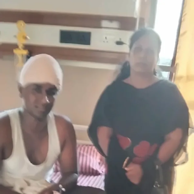A man with a white bandage on his head sitting beside a woman in a black outfit, in a hospital room